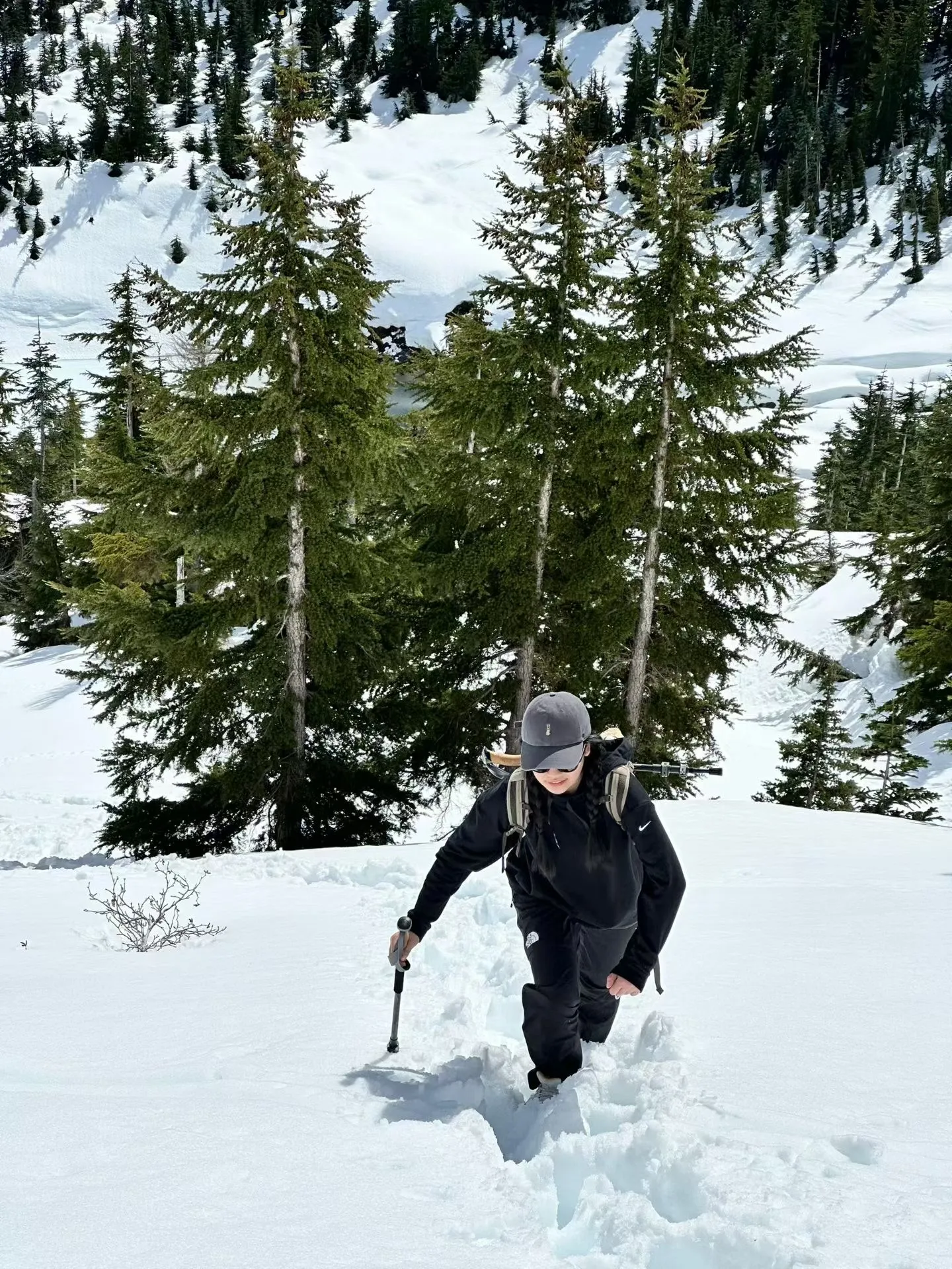 Jieyi Deng hiking uphill through deep snow in a black jacket and cap, with snow-laden evergreens and a mountain slope behind.